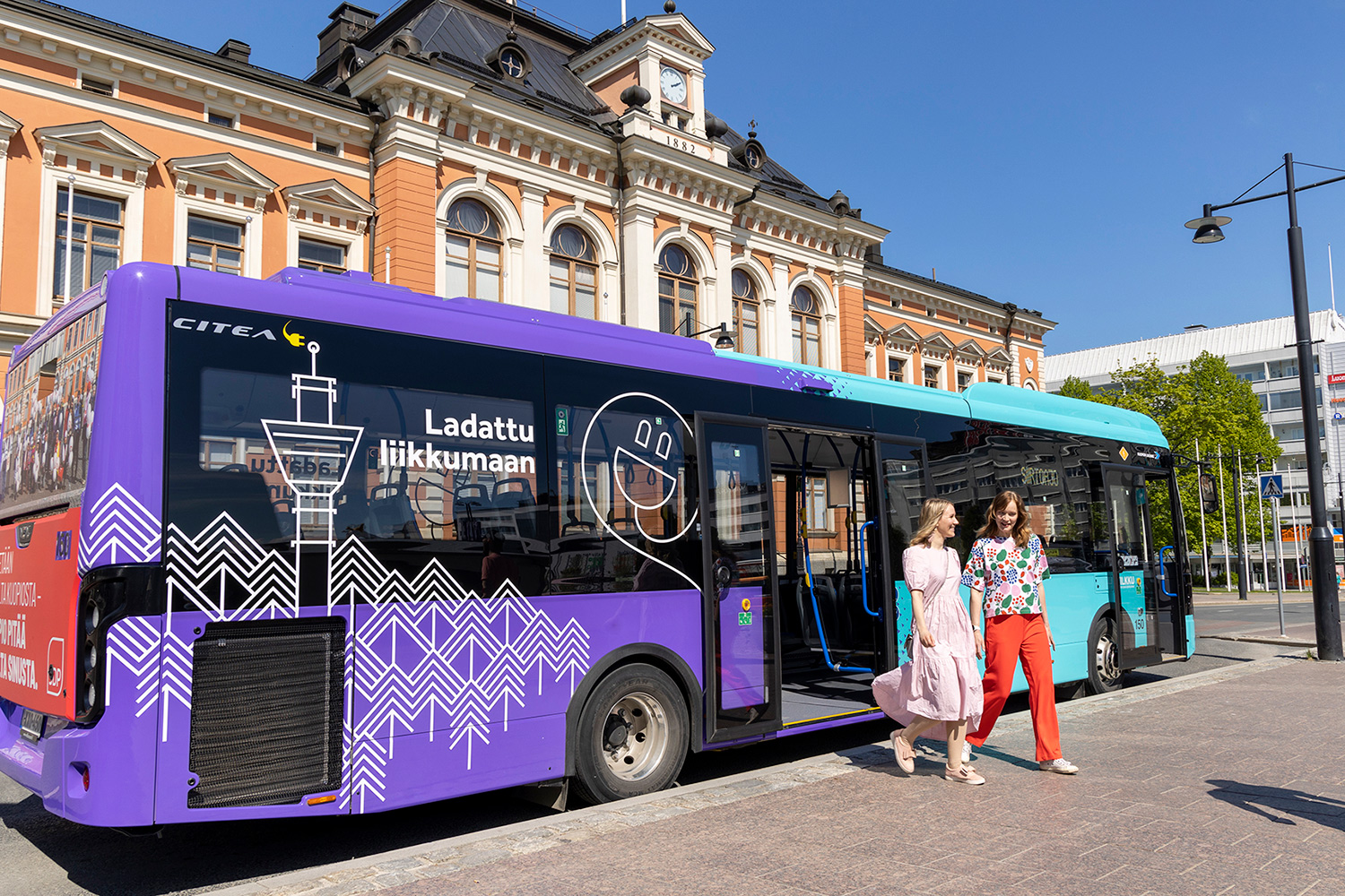 Turkoosi-violetti sähköbussi on pysähtynyt kaupungintalon eteen kauniina kesäpäivänä ja kaksi ystävystä nousee bussista torille.